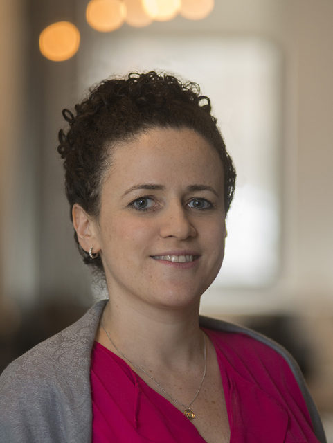 Woman with curly brown hair, wearing a pink top and gray sweater, smiles at the camera. The background is softly blurred, with warm lights and a window visible.