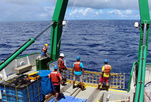 Four people wearing safety gear stand on the deck of a ship at sea, working with equipment and containers under a partly cloudy sky.