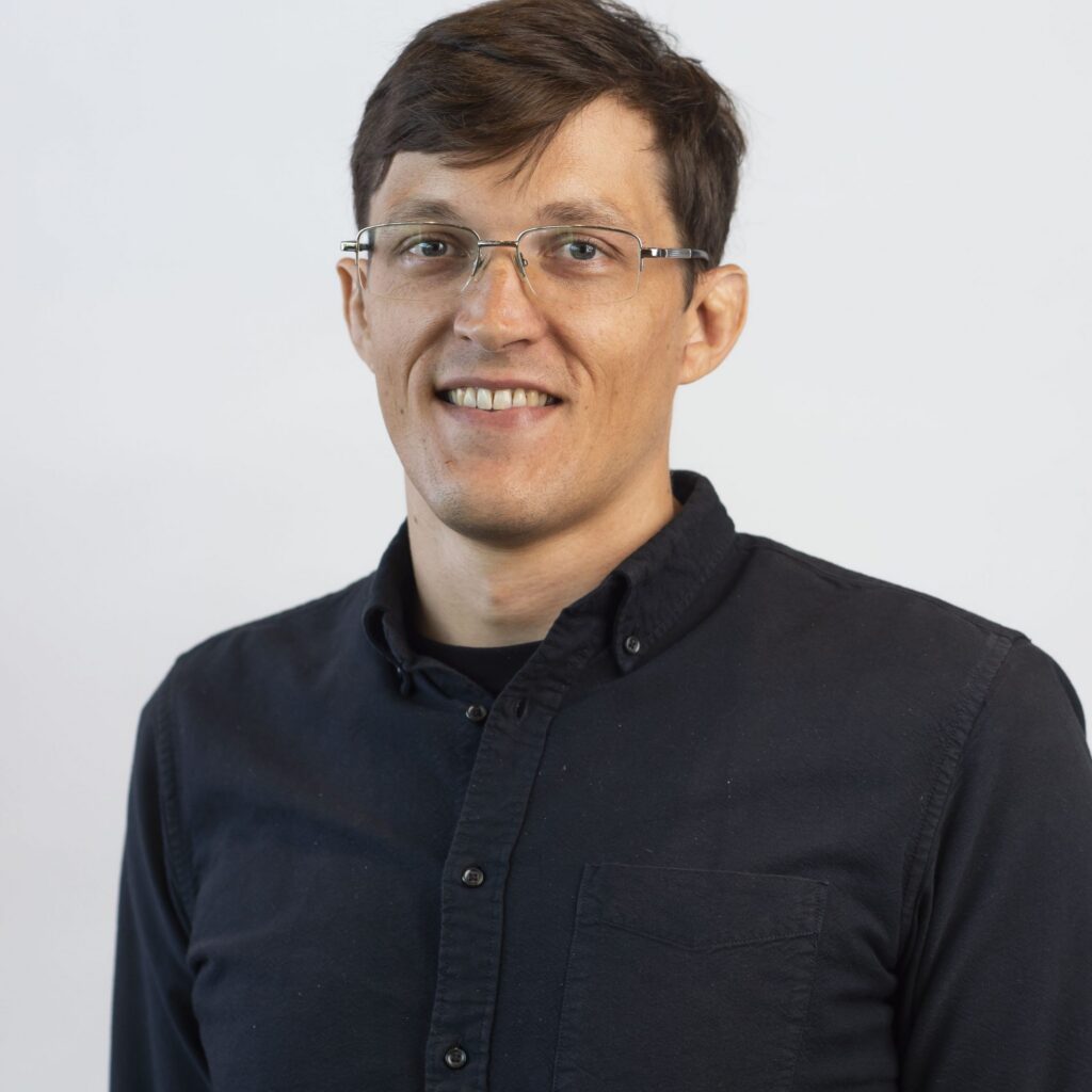 A person with short brown hair, glasses, and a dark button-up shirt smiles at the camera against a plain white background.