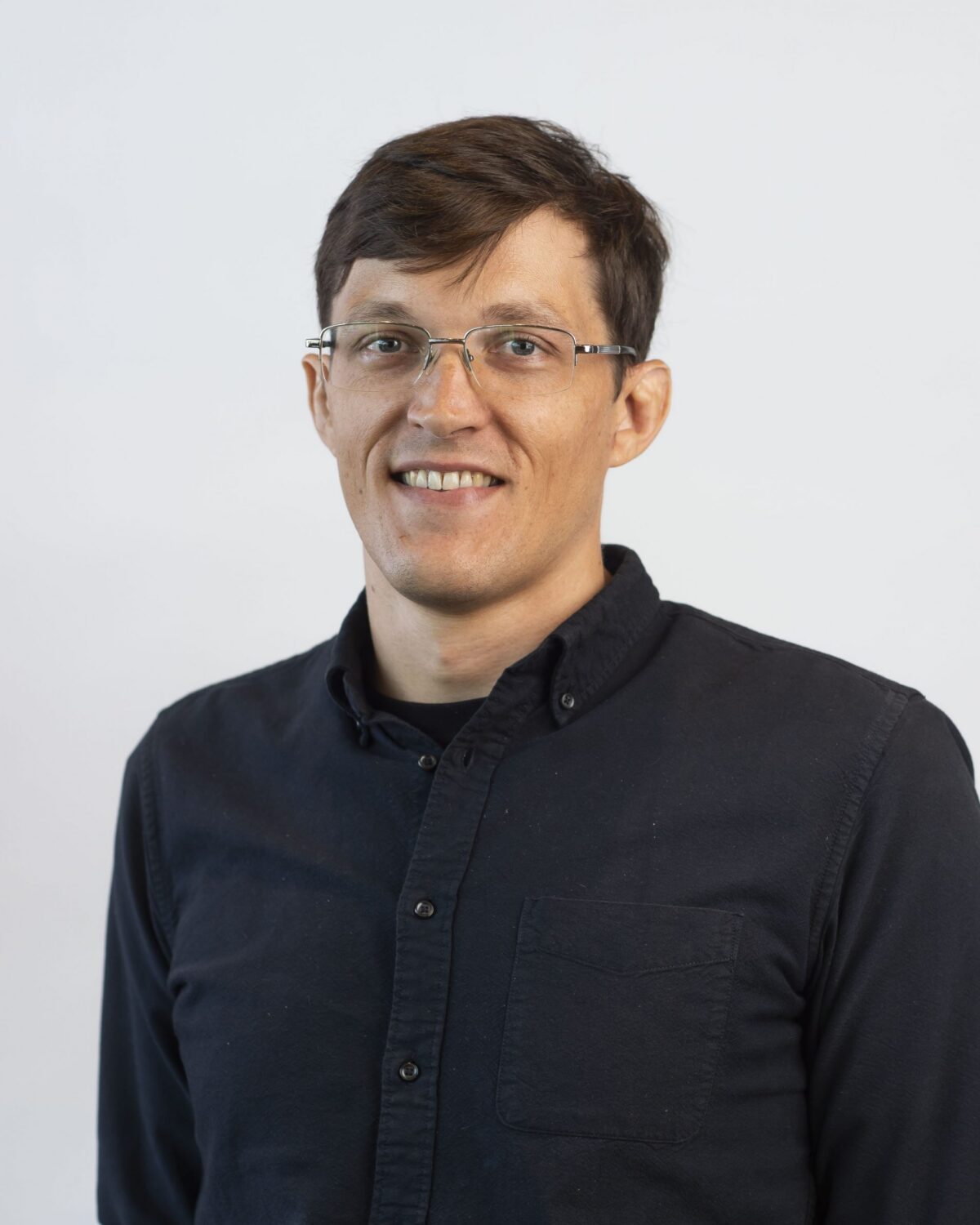 A person with short brown hair, glasses, and a dark button-up shirt smiles at the camera against a plain white background.