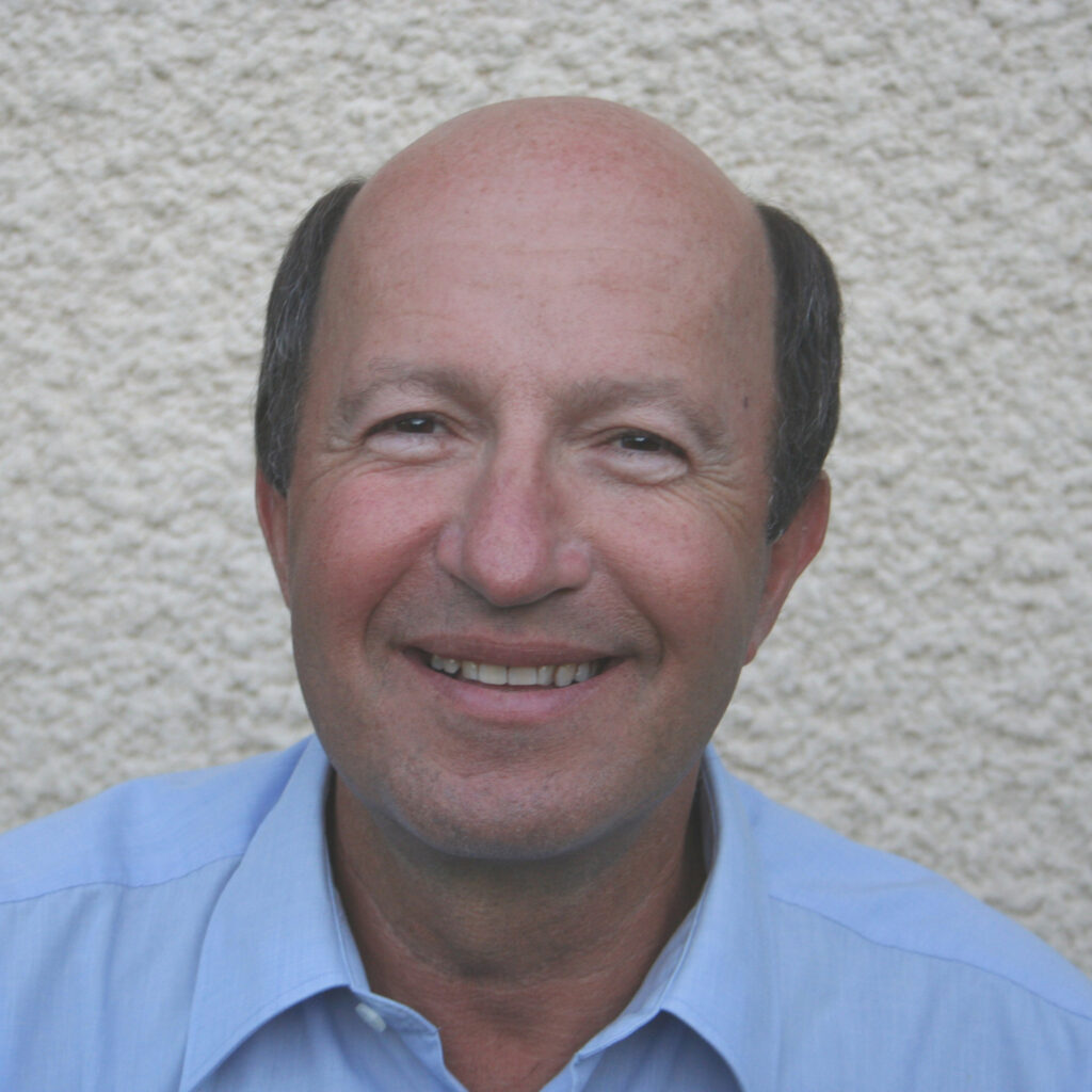 A middle-aged man with a bald head, wearing a light blue collared shirt, smiling in front of a textured off-white wall.