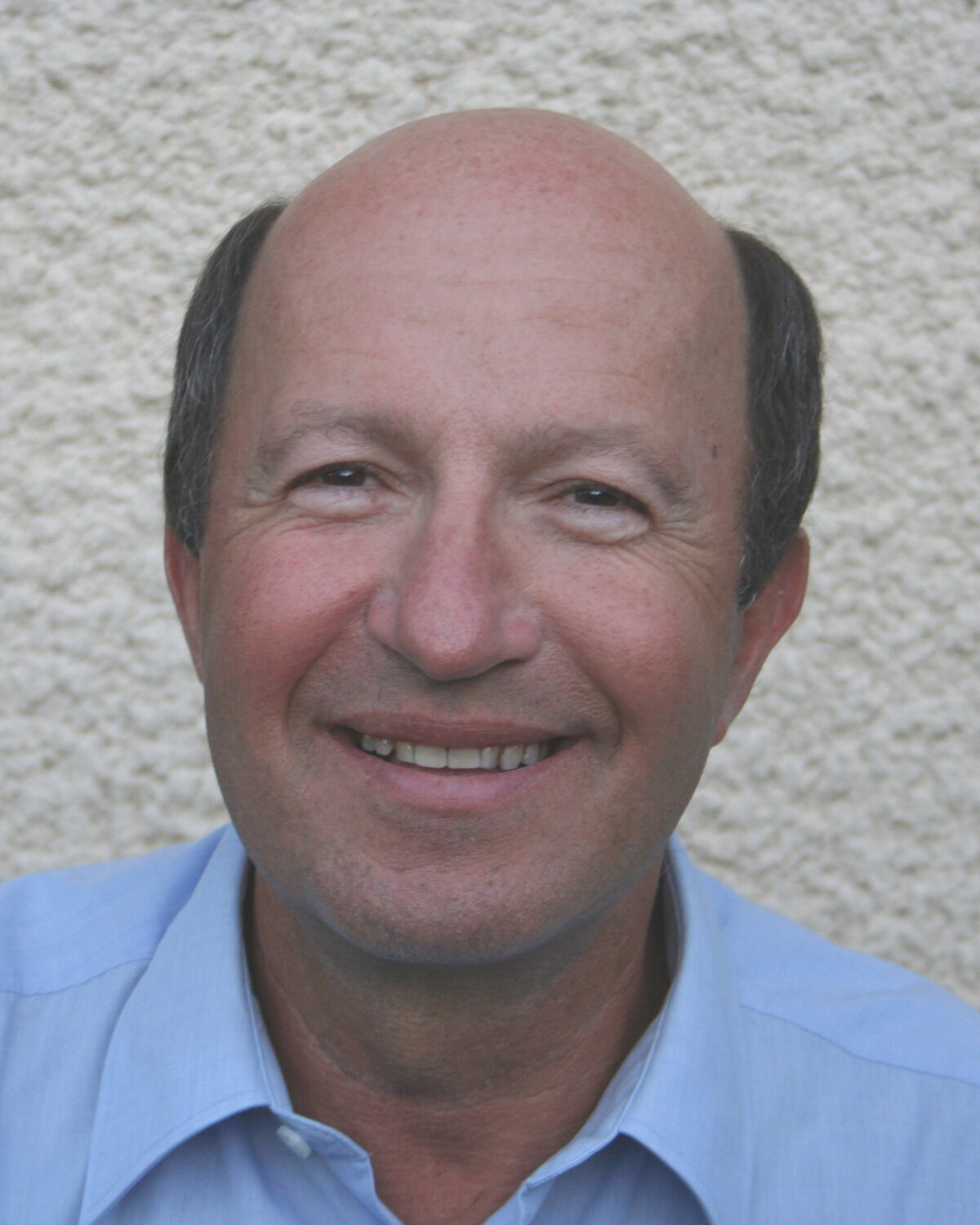 A middle-aged man with a bald head, wearing a light blue collared shirt, smiling in front of a textured off-white wall.