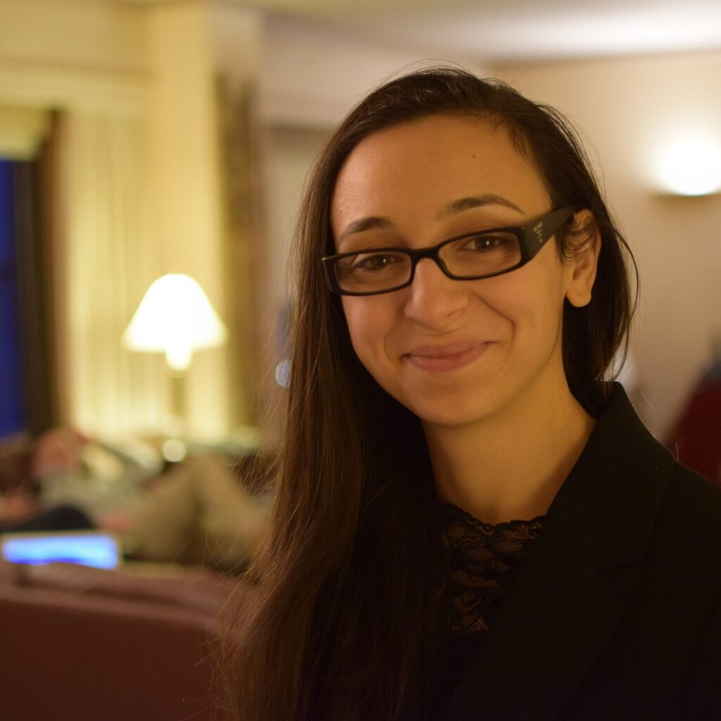 A woman with long dark hair and glasses smiles at the camera indoors, while two people relax on couches in the softly lit background.
