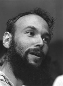 A black and white photo of a bearded man with curly hair, looking slightly upward and smiling, with a blurred background.