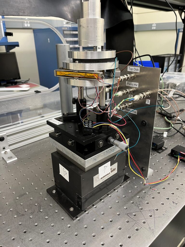 A close-up of a scientific instrument with wires, metal components, and labeled parts, set on a perforated metal table in a laboratory setting.
