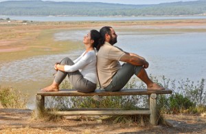 A man and woman sit back to back on a bench overlooking a lake, both looking relaxed and thoughtful, surrounded by nature.