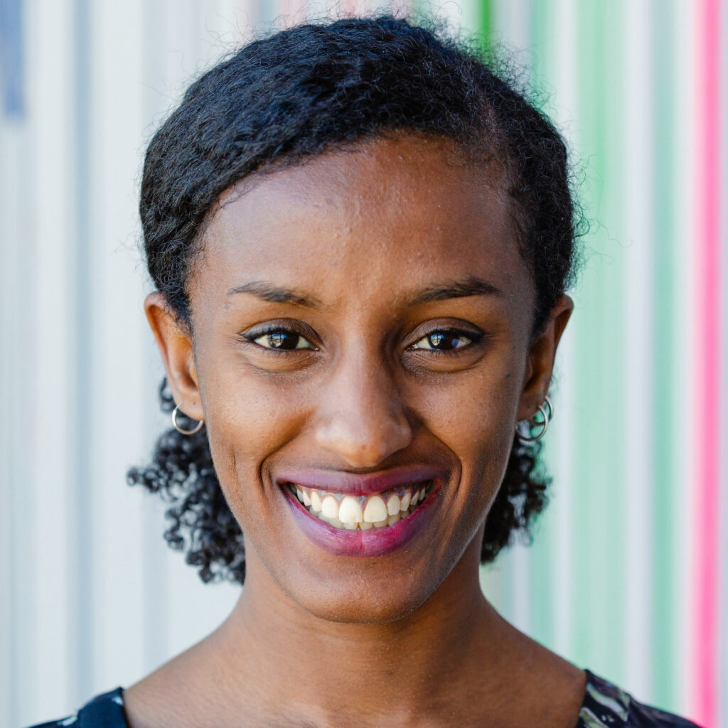 A woman with curly black hair smiles at the camera. She is wearing a black top and hoop earrings, standing in front of a background with vertical colorful stripes.