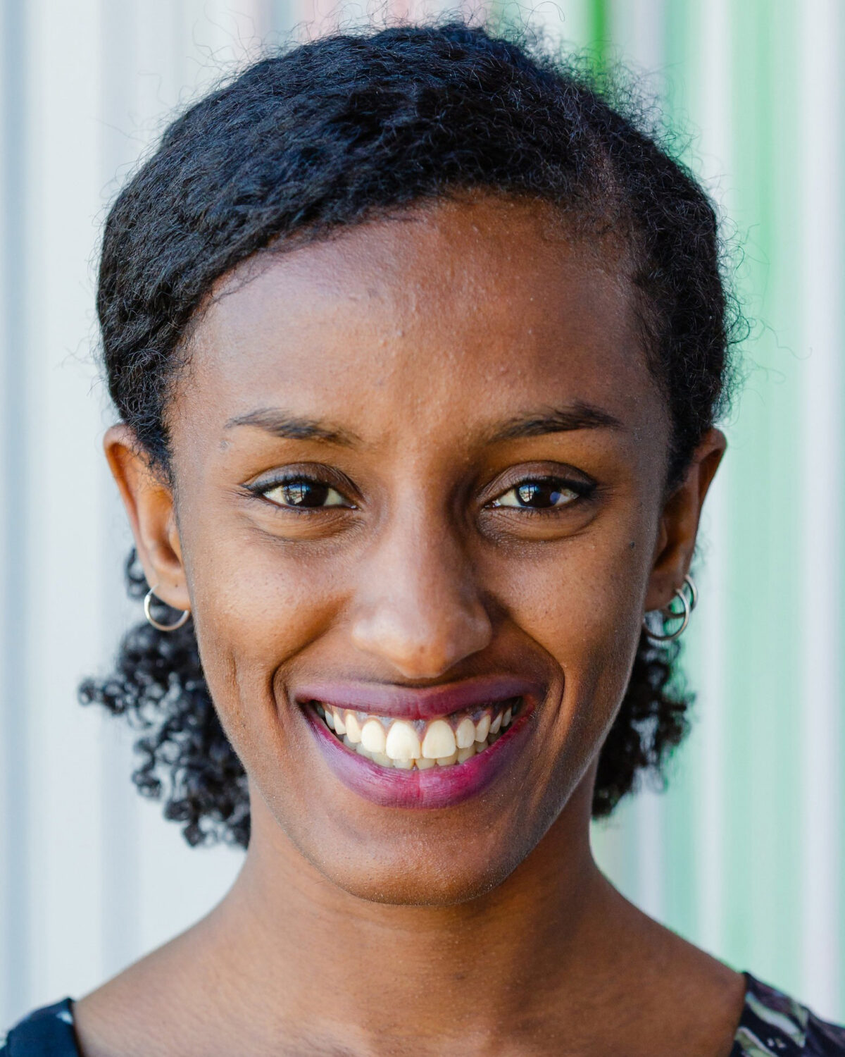 A woman with curly black hair smiles at the camera. She is wearing a black top and hoop earrings, standing in front of a background with vertical colorful stripes.