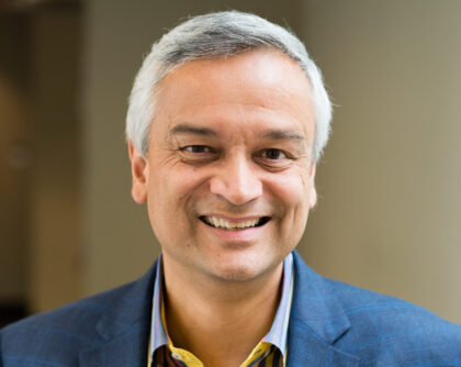Smiling man with short gray hair wearing a blue blazer and a collared shirt, standing indoors with a blurred background.