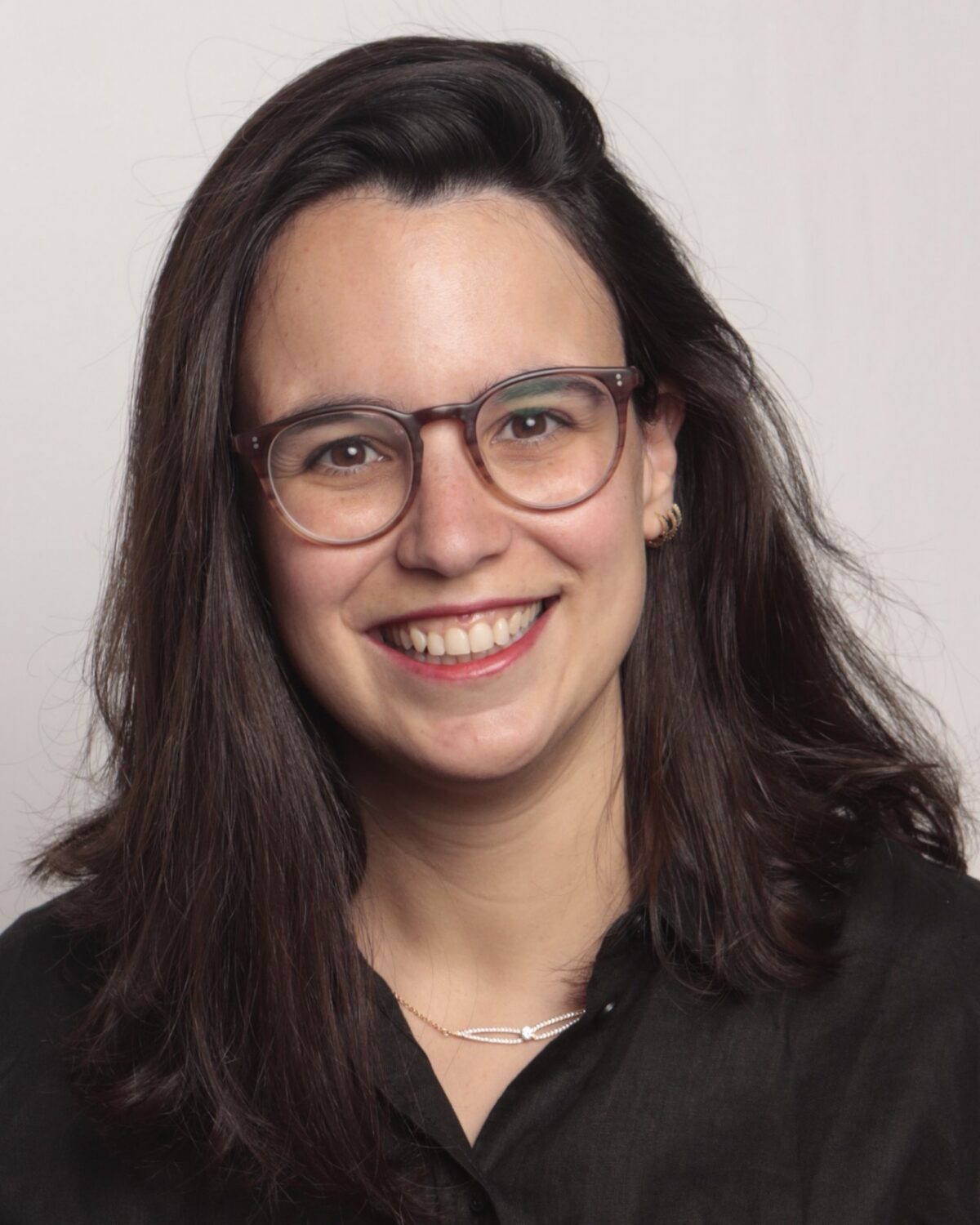 A woman with long dark hair, wearing glasses and a black shirt, smiles at the camera against a plain light background.