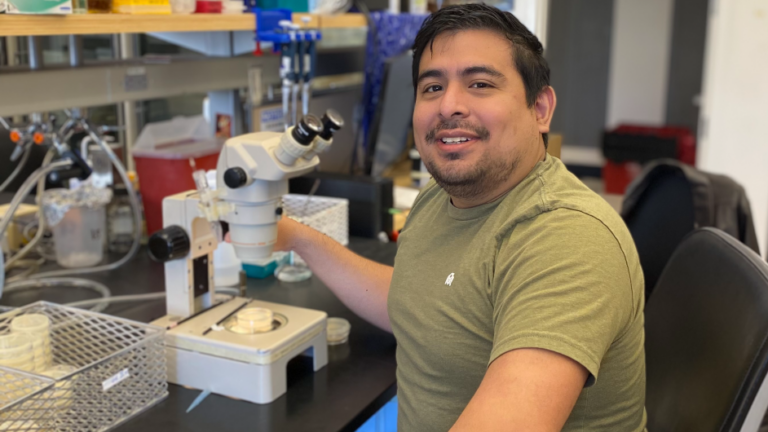A person in a green shirt smiles while sitting at a lab table, using a microscope. Laboratory equipment and supplies are visible on the counter around them.
