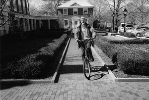 A person rides a bicycle down a brick pathway, smiling, with buildings, bushes, and parked cars visible in the background on a sunny day.