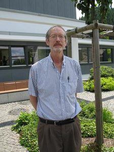 A man with glasses, a mustache, and a striped short-sleeve shirt stands outdoors on a cobblestone path with greenery and a building in the background.