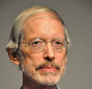 An older man with light skin, gray hair, a mustache, and a beard is wearing glasses and looking into the camera with a neutral expression. The background is plain and gray.