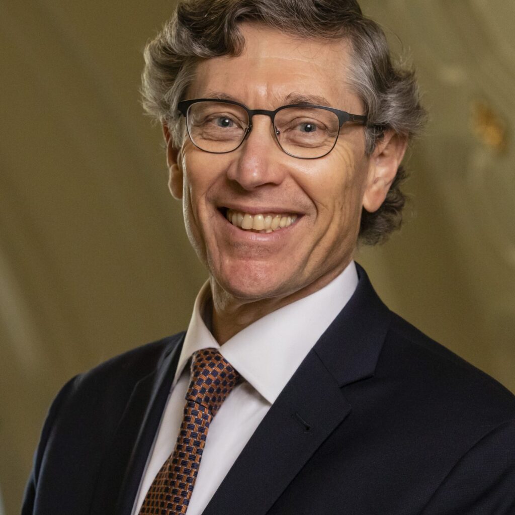 A man with wavy gray hair, glasses, and a dark suit with a white shirt and patterned tie smiles at the camera. The background is softly lit and elegant.