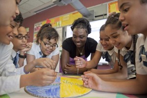 A group of students gathers around a table, smiling and working together on a colorful circular project, while a teacher watches and encourages them. Classroom posters and bright decor are visible in the background.