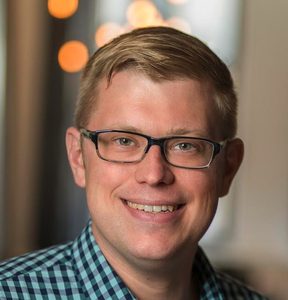 Smiling person with short blond hair and glasses, wearing a blue and black checkered shirt, stands indoors with soft, blurry lights in the background.