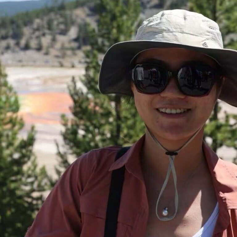A person in sunglasses and a hat smiles in front of the Grand Prismatic Spring at Yellowstone, with green trees and blue sky in the background.