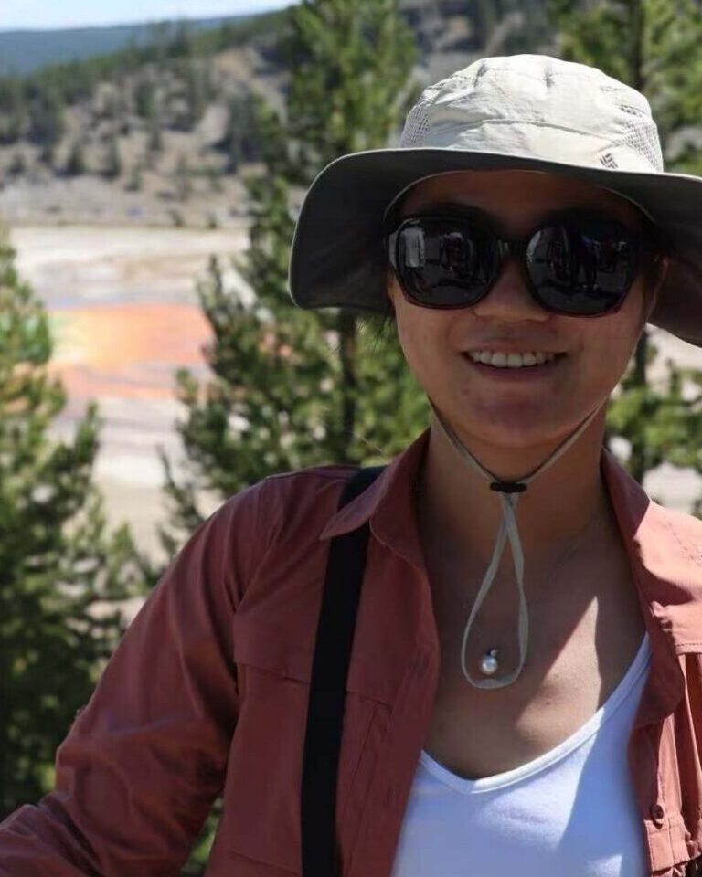 A person in sunglasses and a hat smiles in front of the Grand Prismatic Spring at Yellowstone, with green trees and blue sky in the background.