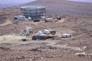 A remote scientific research station sits on rocky terrain, featuring a large circular structure, satellite dishes, small buildings, vehicles, and equipment spread across a barren landscape under a clear sky.