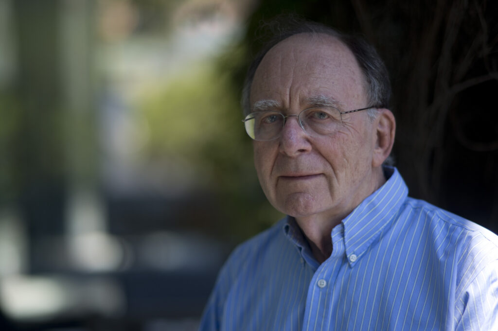 An older man with glasses wearing a blue striped shirt stands outdoors, looking at the camera with a neutral expression. The background is blurred with greenery.