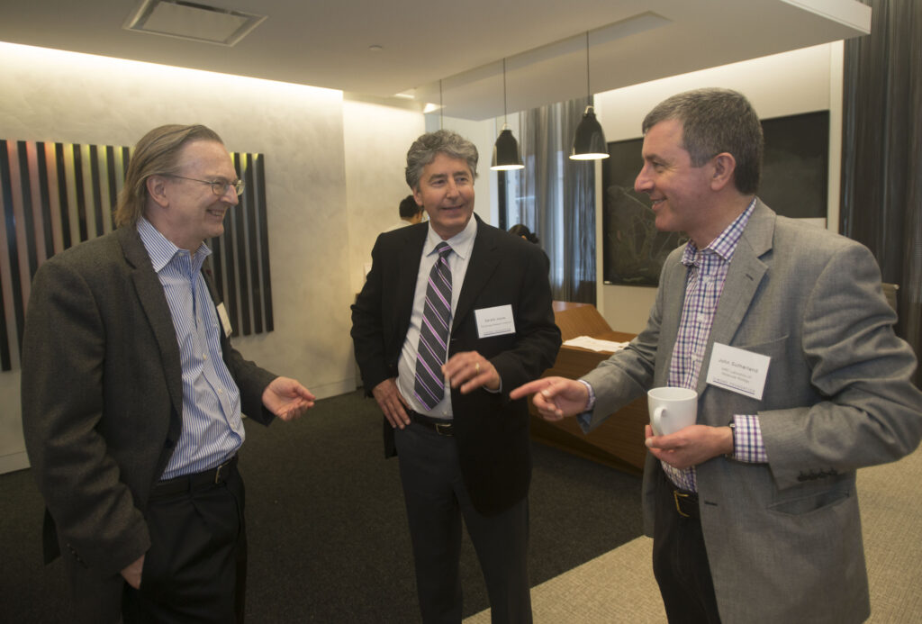 Three men in business attire stand in an office, smiling and talking. Two wear nametags and one holds a cup. Striped artwork hangs on the wall behind them.