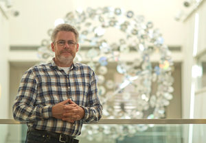 A man wearing glasses and a plaid shirt stands indoors, leaning on a glass railing with a large modern sculpture made of circular elements blurred in the background.
