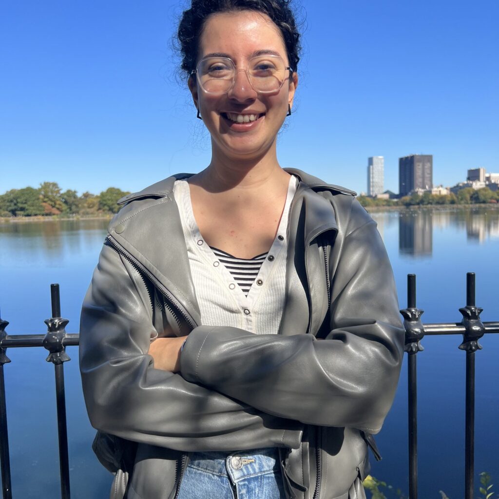 A person with curly hair, glasses, and a gray jacket smiles with arms crossed in front of a lake and black fence, with buildings and trees visible in the background under a clear blue sky.