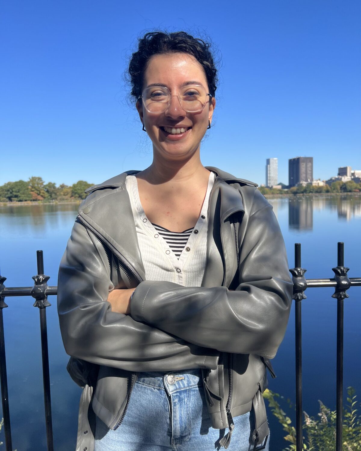 A person with curly hair, glasses, and a gray jacket smiles with arms crossed in front of a lake and black fence, with buildings and trees visible in the background under a clear blue sky.