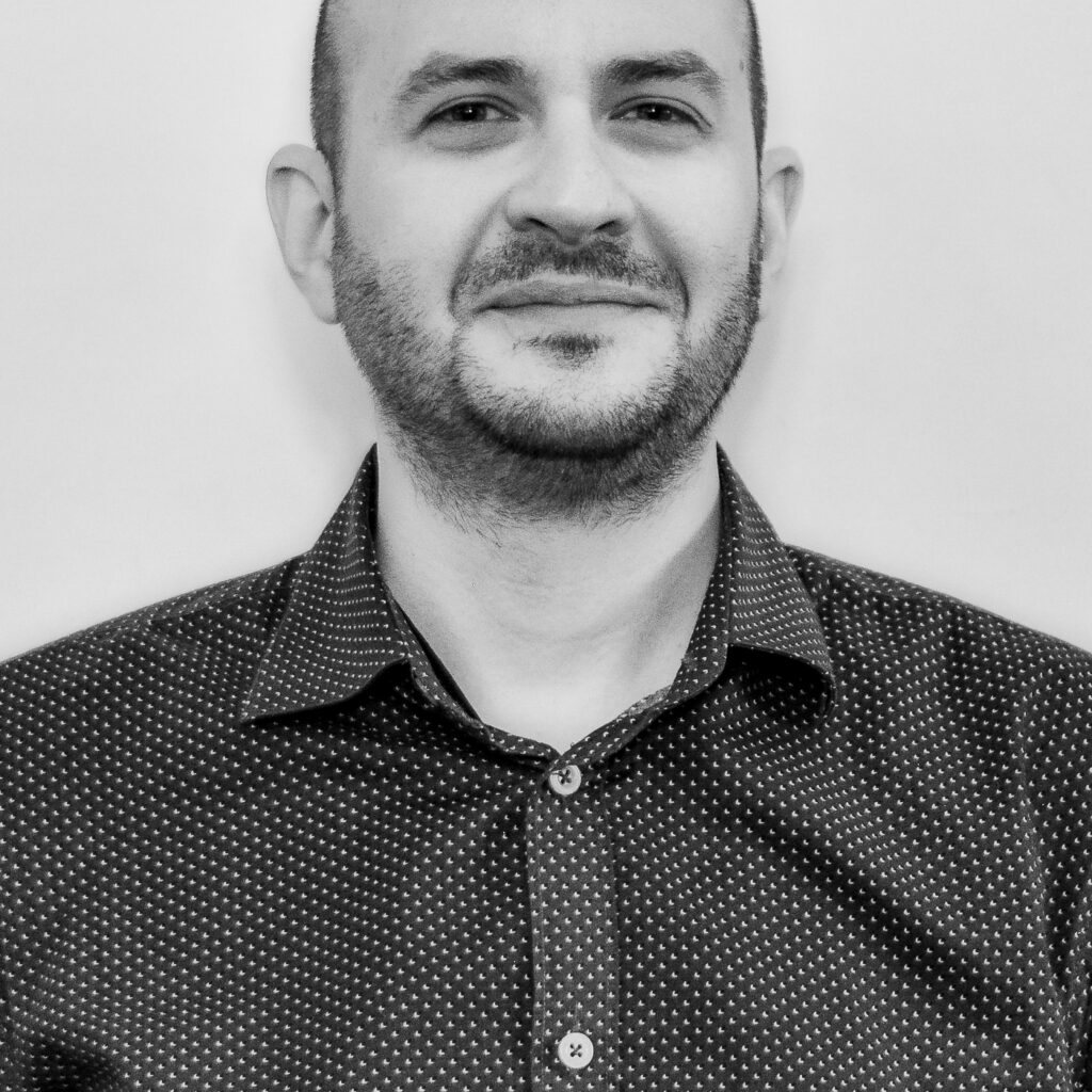 A man with a shaved head and beard, wearing a dark, patterned button-up shirt, stands against a plain light background, looking at the camera and smiling slightly. Black and white photo.