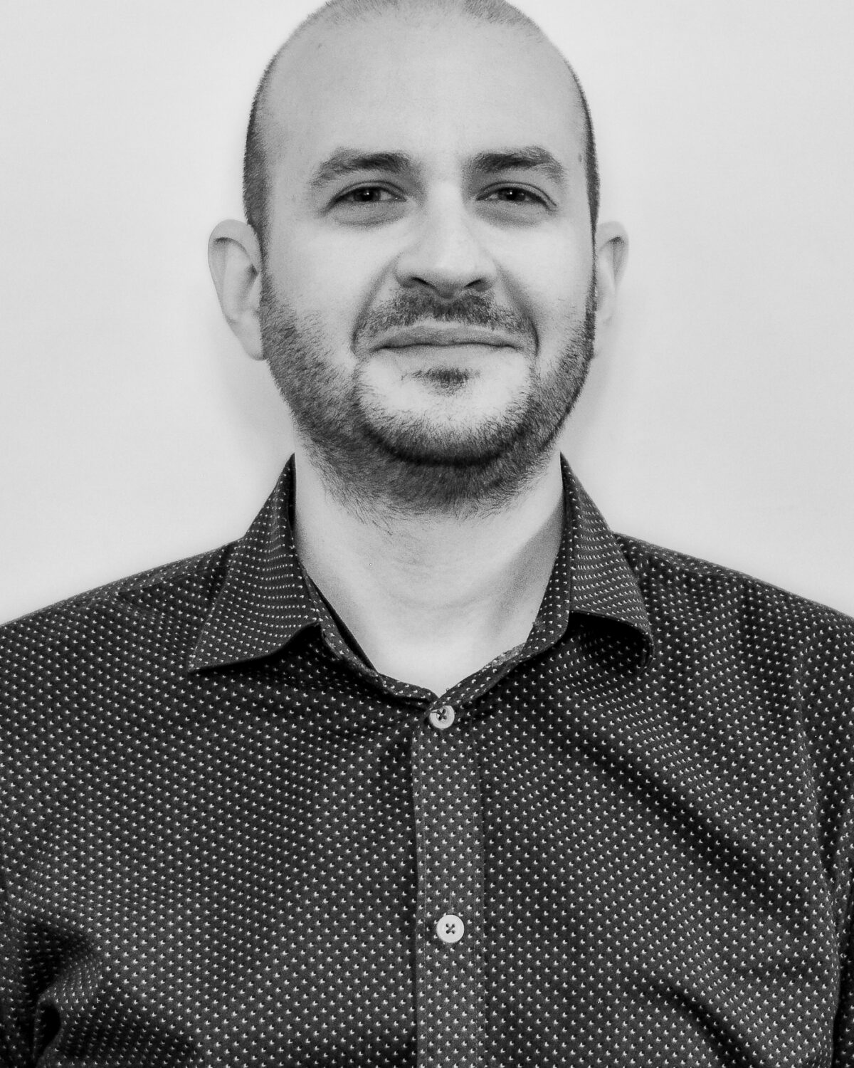 A man with a shaved head and beard, wearing a dark, patterned button-up shirt, stands against a plain light background, looking at the camera and smiling slightly. Black and white photo.
