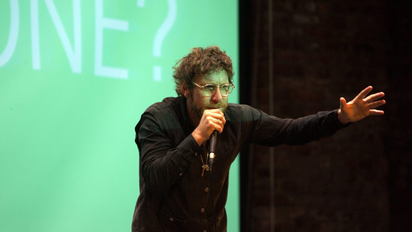 Man standing on a stage and leaning forward with an arm outstretched while the other holds a microphone to his mouth