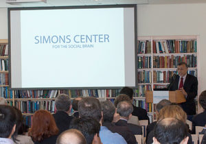 A speaker addresses an audience in a library-like room, standing at a podium next to a screen displaying Simons Center for the Social Brain..