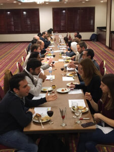 A group of people sit on both sides of a long table in a conference room, eating food and engaging in conversation. Plates, glasses, and papers are visible on the table.