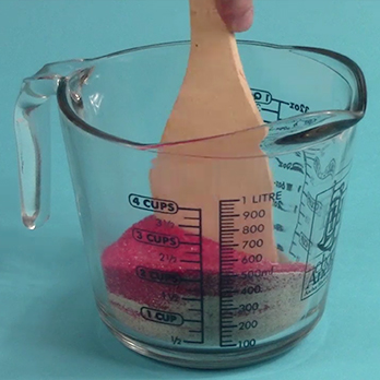 Measuring cup with pink sand inside it and a wooden spoon in the sand