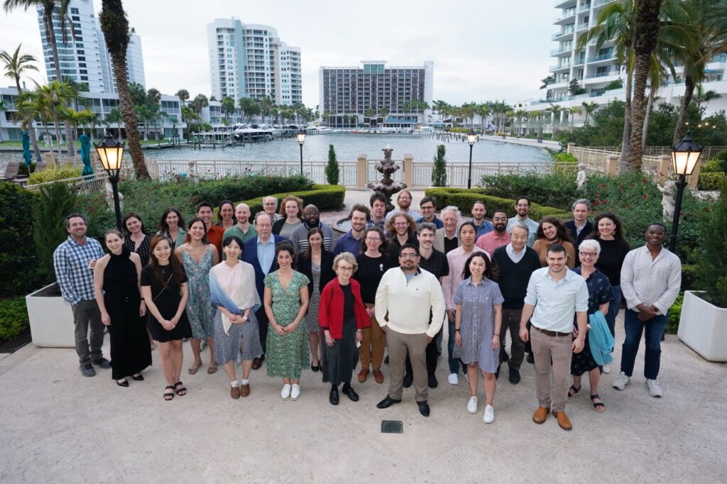 A group of around 40 people pose together outdoors in front of a marina with tall buildings, palm trees, and gardens, all smiling at the camera.