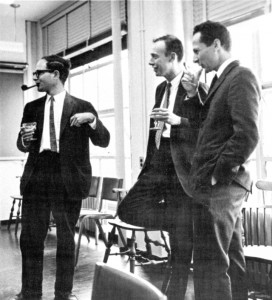 Three men in suits stand in a room with chairs and large windows, talking and holding drinks. One gestures with his hand while the others listen and smile. The photo is black and white.