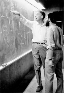 Two men stand at a chalkboard covered in equations and diagrams; one man gestures towards a part of the board while explaining something to the other, who listens attentively.