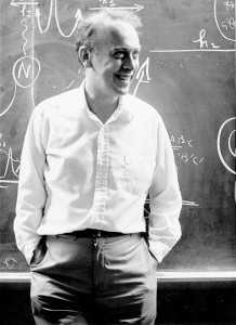A man in a white shirt stands smiling in front of a chalkboard covered with scientific diagrams and equations.