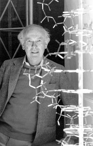 An older man in a suit stands beside a large model of a DNA double helix structure, smiling at the camera. The background includes geometric framework and bright lighting.
