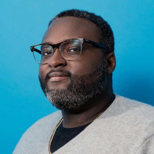 A man with short, curly hair, a beard, and glasses wears a light gray sweater and gold necklace, posing against a plain blue background.