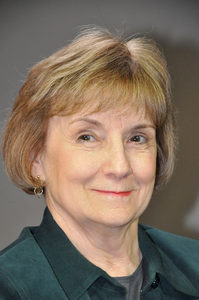 A woman with short, light brown hair and a slight smile is wearing a dark green collared shirt. The background is a plain, neutral color.