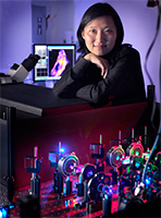 A woman stands behind a table with scientific equipment and colorful lights. A microscope and computer screen displaying an image are visible in the background.