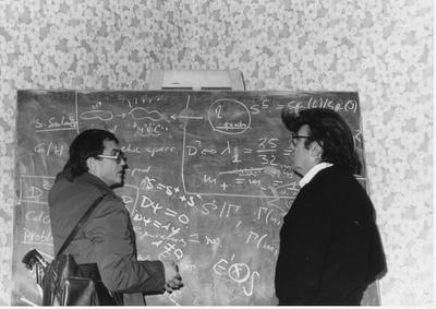 Two people stand in front of a chalkboard covered in mathematical equations, deep in conversation. One wears glasses and carries a bag; the other also wears glasses. The background features patterned wallpaper.