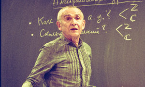 An older man stands in front of a chalkboard with handwritten notes and diagrams. He wears a striped shirt and looks forward, possibly teaching or explaining something. The writing on the board appears to be in Russian.