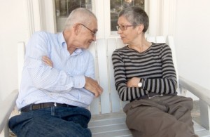 Two older adults sit on a white bench, facing each other with arms crossed, smiling and leaning in as if sharing a lighthearted moment.