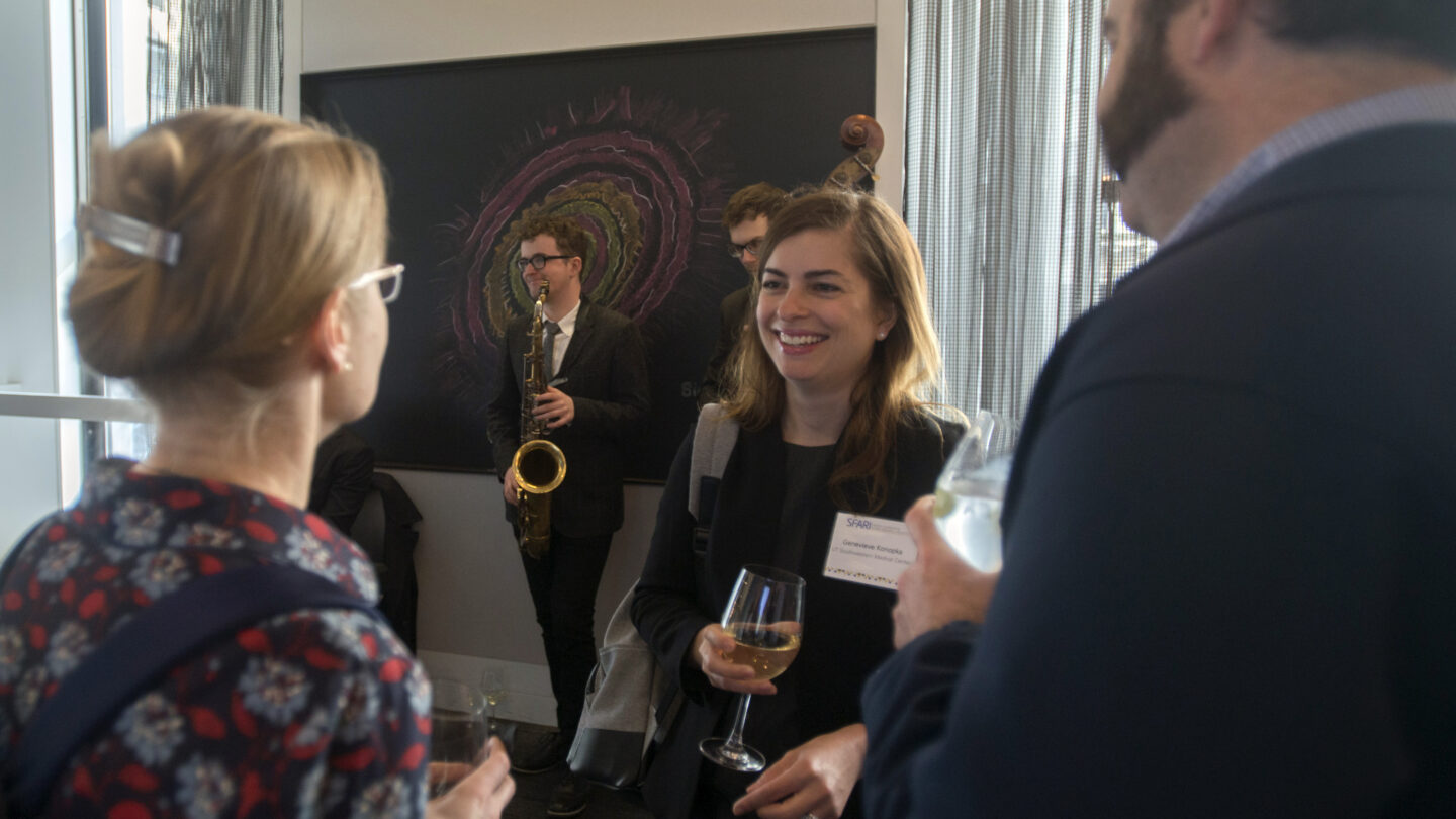 Three people converse at an indoor event, holding drinks. A jazz band plays in the background. One woman smiles while others listen. Abstract art hangs on the wall.