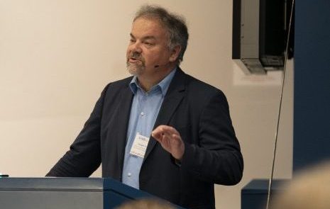 A man in a suit speaks at a podium, wearing a headset microphone and a conference name badge, gesturing with one hand while delivering a presentation.