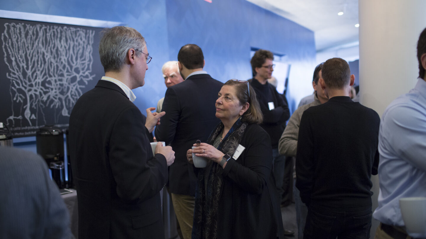 People in business attire stand and converse, holding coffee cups, at a professional networking event in a modern, blue-walled room. A scientific diagram is visible on the wall in the background.