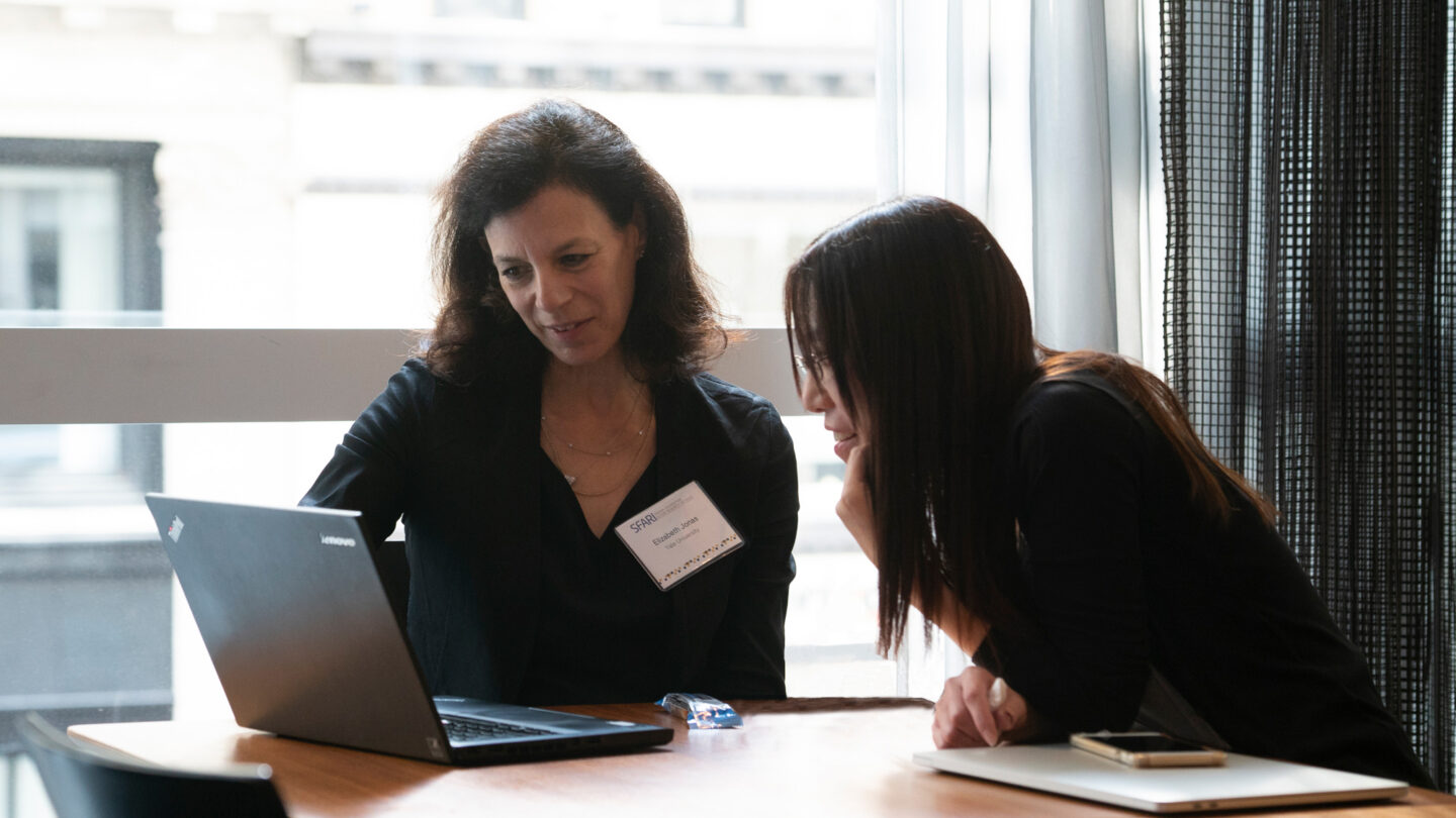 Two women sit at a table, looking at a laptop. One points at the screen while the other observes closely. They both appear focused and engaged, with notebooks and a mouse on the table near them.